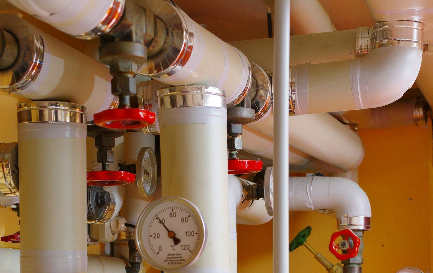 Insulated heating pipes, red valve handles, and temperature gauges on a yellow wall.