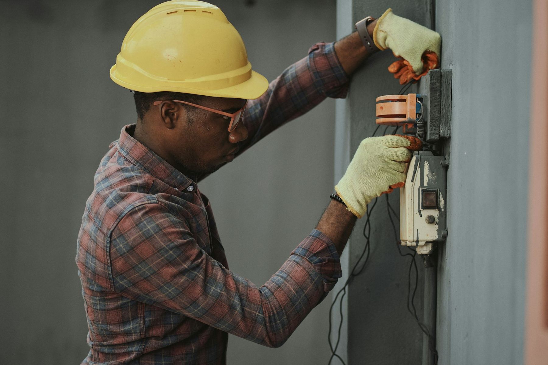 A technician in a hard hat and gloves working on a wall-mounted control box.